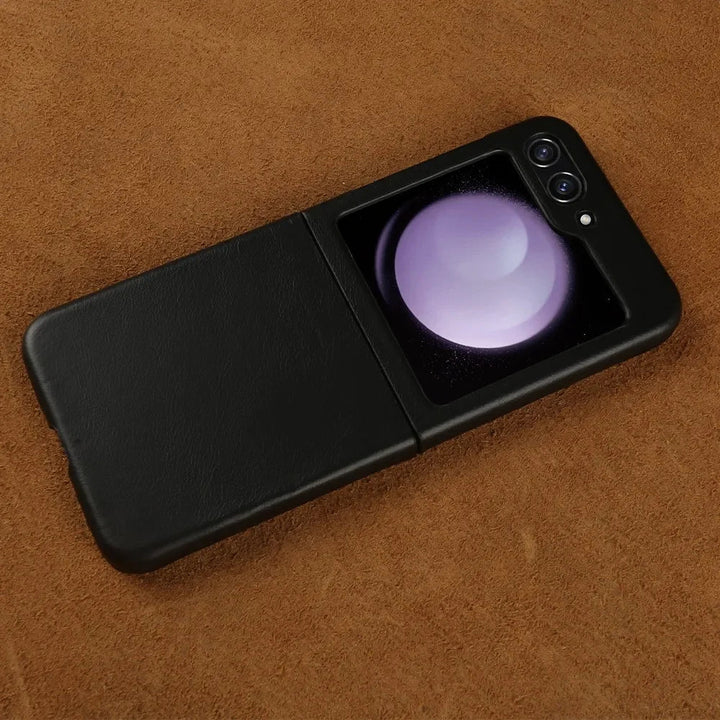 A black Samsung Galaxy Z Flip foldable smartphone with dual rear cameras is partially open, resting on a brown textured surface. The external display shows a circular, glowing graphic. Features shockproof protection for added durability.