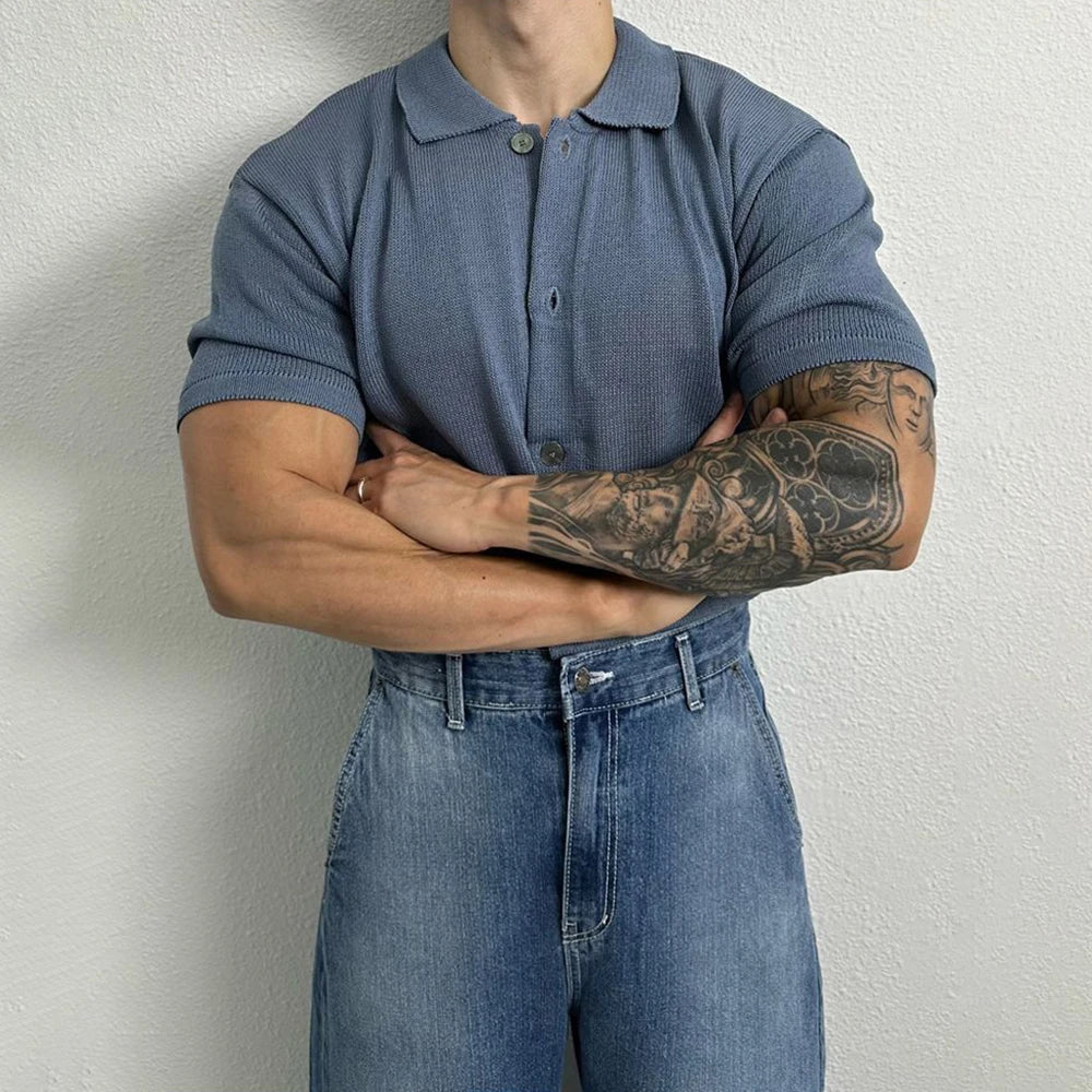 A person stands against a light textured wall with arms crossed, wearing a short-sleeve unisex top in blue and blue jeans. One arm features a detailed black tattoo sleeve. The persons face is not visible.