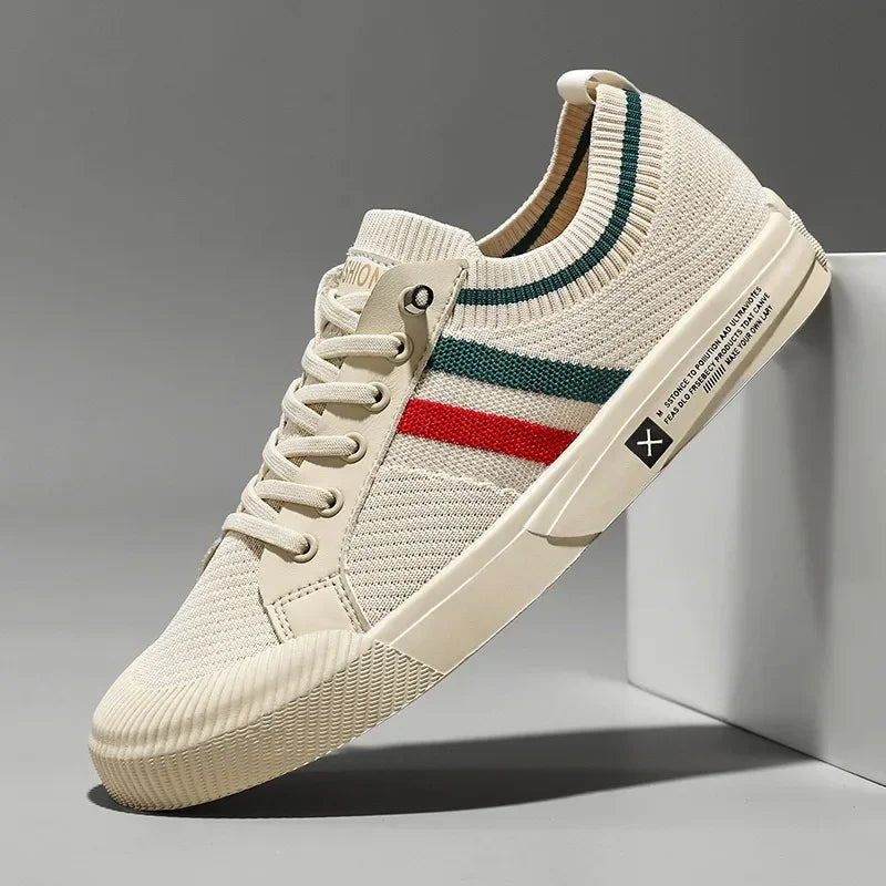 A beige knit sneaker with a breathable mesh upper, cream outsole, beige laces, and green and red stripes on the side. The shoe is angled against a white block on a gray background.