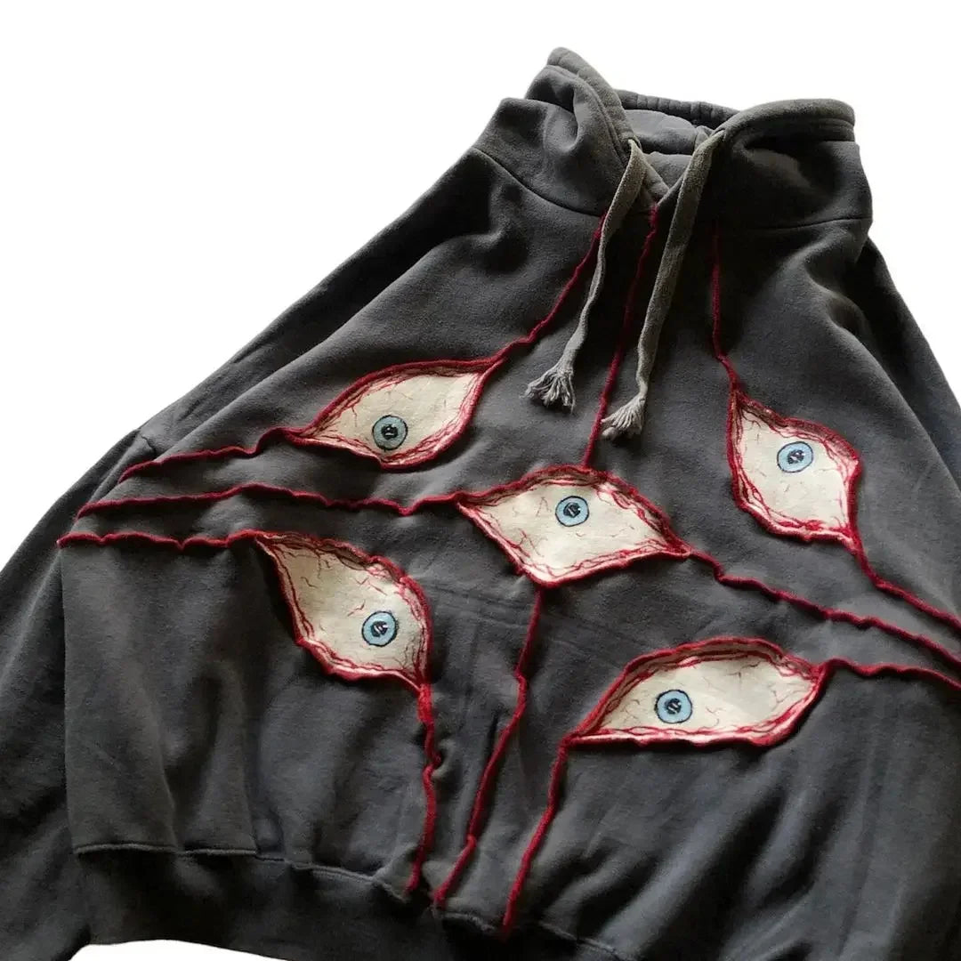 A dark gray hoodie with an eye pattern patchwork—five eye-shaped fabric patches on the front, each outlined in red stitching with a blue eyeball. Made from breathable fabric, it features a drawstring hood and is laid flat.