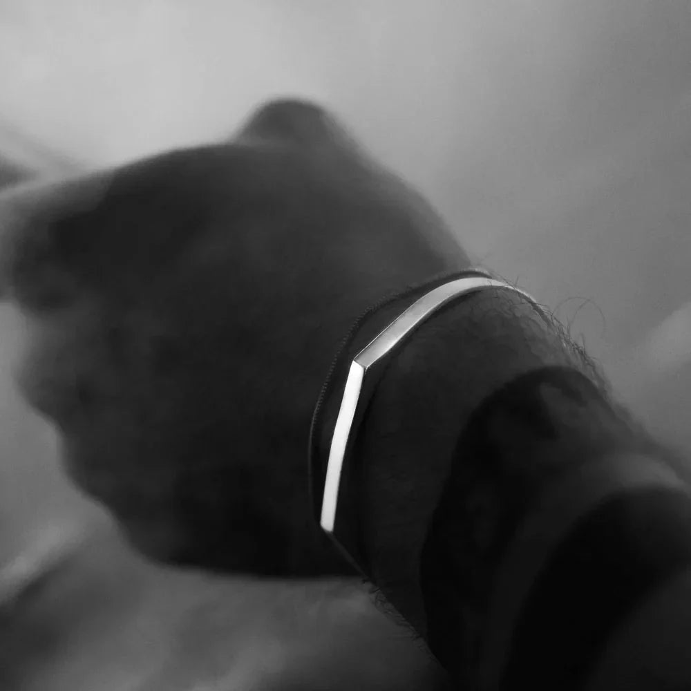 A close-up, black and white photo of a persons wrist wearing a slim, minimalist cuff with a subtle V-shaped bend. The stainless steel bracelet stands out against the blurred background.