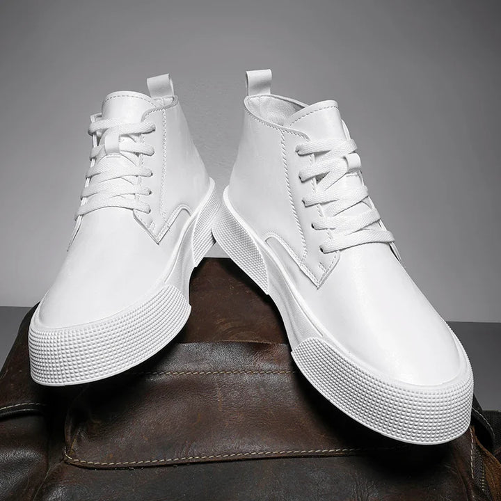 A pair of vintage-inspired high-tops in white with thick soles, laces, and smooth finish, featuring traditional craftsmanship, displayed on a brown leather surface against a gray background.