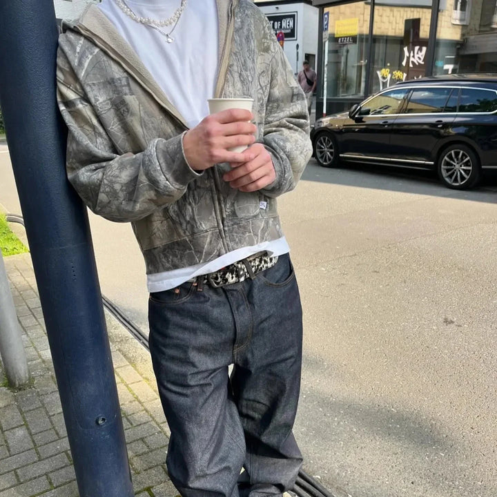 A person in an oversized fit camouflage hoodie, white t-shirt, baggy jeans, and a beaded belt leans against a pole on a city sidewalk, holding a white cup. A black car is parked on the street in the background.