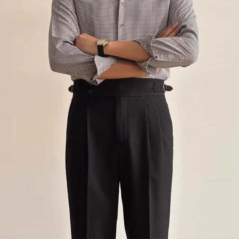 A person wearing a black and white checkered shirt with sleeves rolled up, luxury tailored pants, and a black wristwatch stands with arms crossed against a plain background.