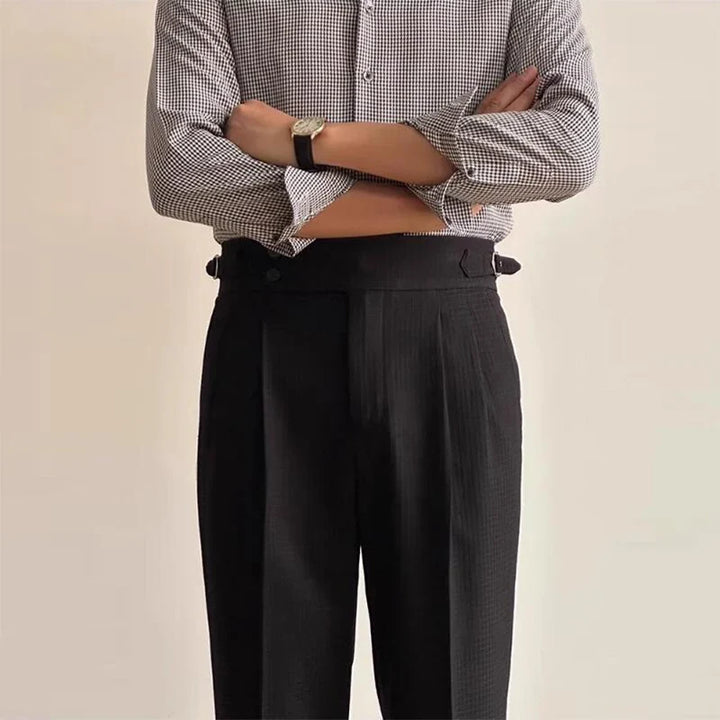 A person wearing a black and white checkered shirt with sleeves rolled up, luxury tailored pants, and a black wristwatch stands with arms crossed against a plain background.