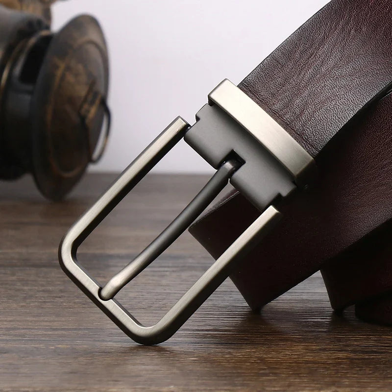 A close-up of a brown cowhide belt with a brushed metal buckle resting on a wooden surface, with a blurred background featuring a pair of headphones.