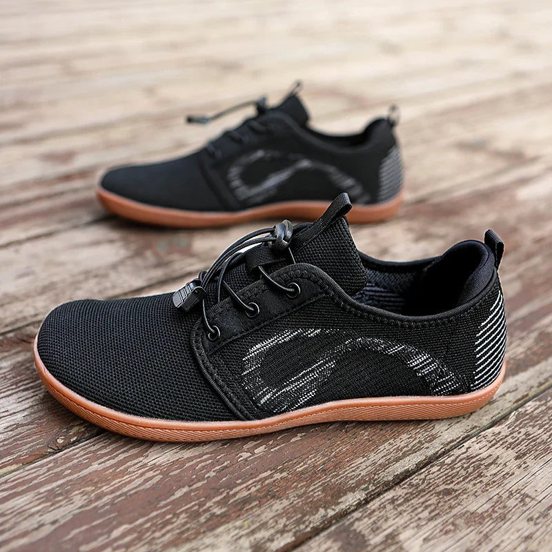 A pair of black athletic sneakers with brown soles and white accents, placed on a rustic wooden surface. Featuring mesh fabric, elastic laces, and a subtle wave design, these shoes offer a cushioned sole for all-day comfort.