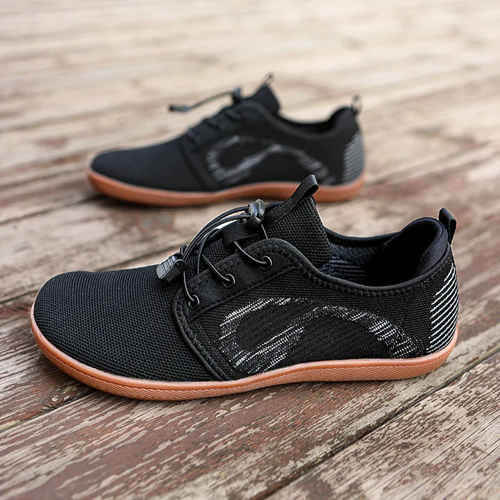 A pair of black athletic sneakers with brown soles and white accents, placed on a rustic wooden surface. Featuring mesh fabric, elastic laces, and a subtle wave design, these shoes offer a cushioned sole for all-day comfort.