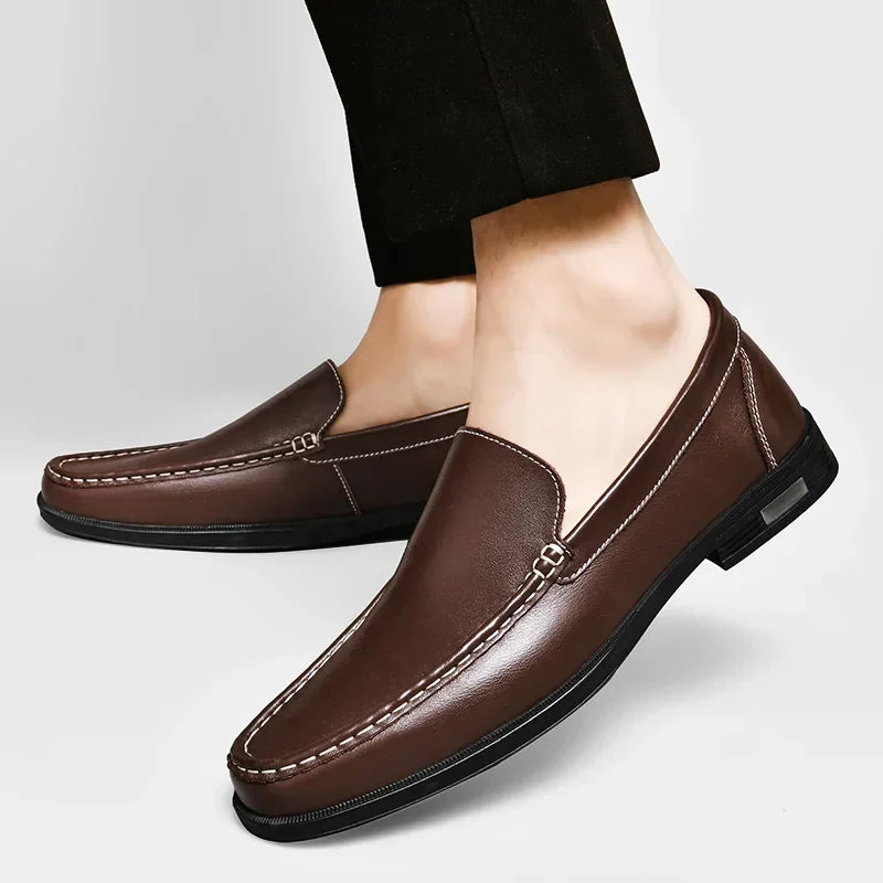 A person wearing classic slip-on loafers crafted from genuine leather, featuring white stitching and black soles, paired with black pants, against a plain light gray background.
