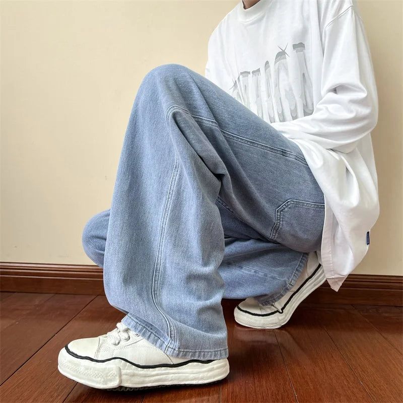 A person wearing relaxed fit, light blue jeans, a white graphic t-shirt, and white sneakers is squatting on a wooden floor against a beige wall. The persons face is not visible.