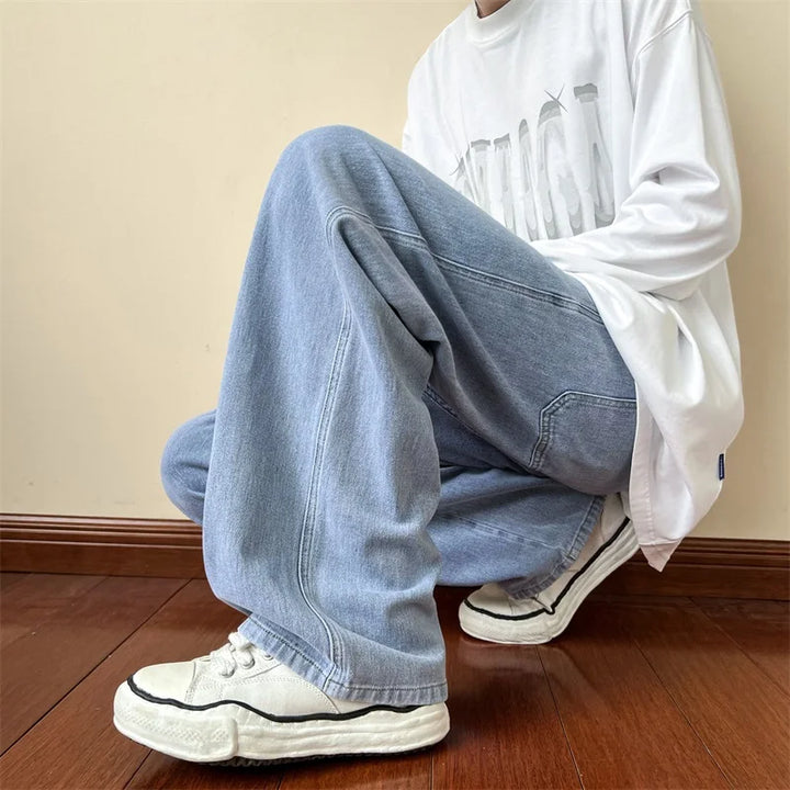 A person wearing relaxed fit, light blue jeans, a white graphic t-shirt, and white sneakers is squatting on a wooden floor against a beige wall. The persons face is not visible.