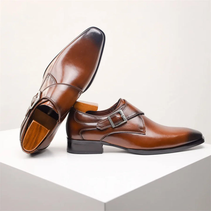 A pair of polished brown Italian leather monk strap shoes with a single silver buckle, displayed on a white geometric surface. One handcrafted shoe is upright while the other rests with its sole visible.