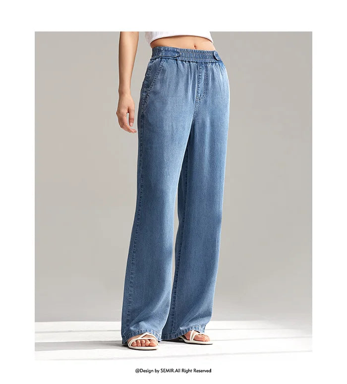 A person wearing loose-fitting, wide-leg, light blue denim pants with an elastic waistband and white sandals stands on a light-colored surface against a plain, neutral background.