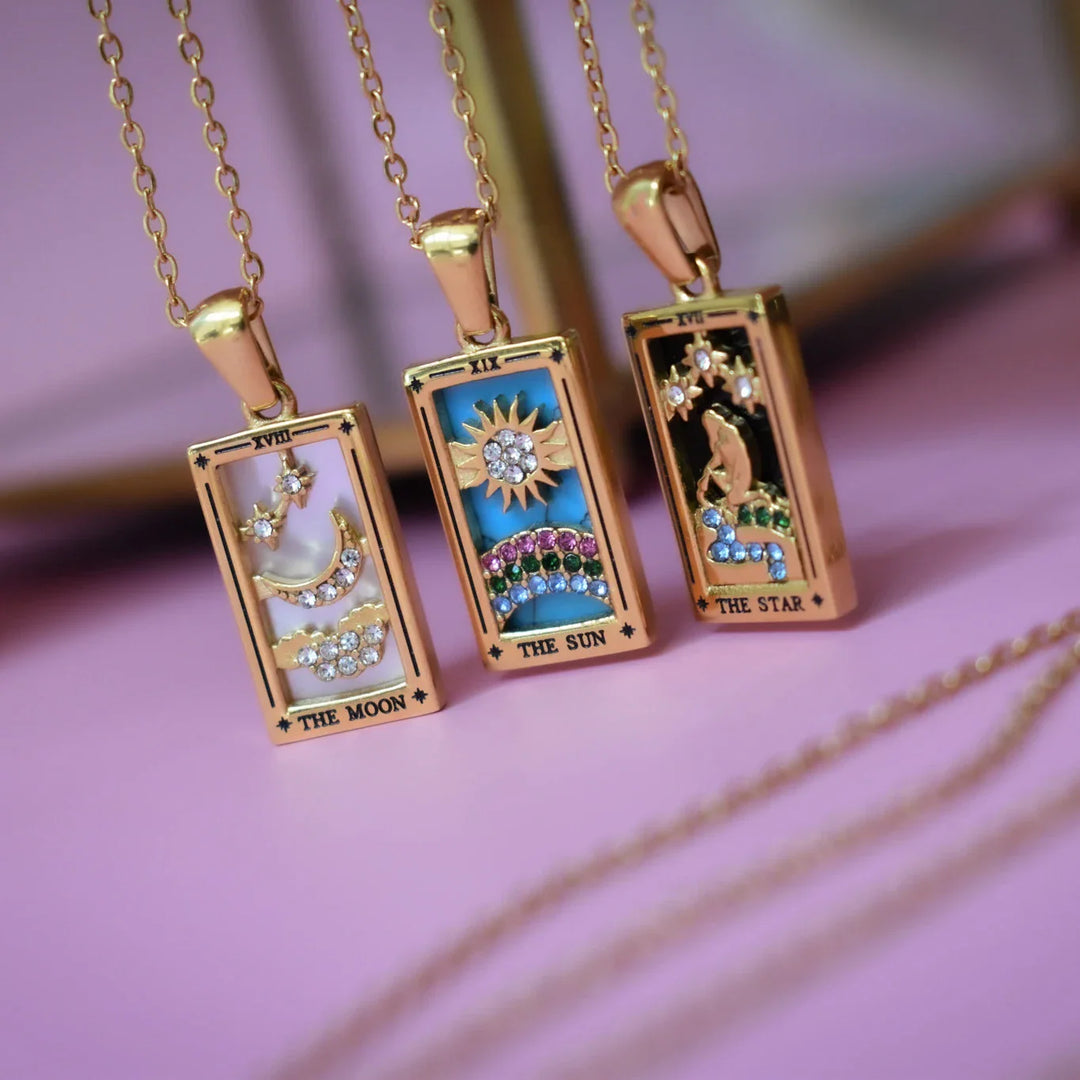 Three 18K gold-plated rectangular pendant necklaces with colorful tarot card designs: one with blue enamel moon and stars, one radiant sun, and one woman by water under stars, labeled The Moon, The Sun, and The Star—our Solaris Pendant Necklace set.