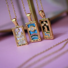 Three 18K gold-plated rectangular pendant necklaces with colorful tarot card designs: one with blue enamel moon and stars, one radiant sun, and one woman by water under stars, labeled The Moon, The Sun, and The Star—our Solaris Pendant Necklace set.