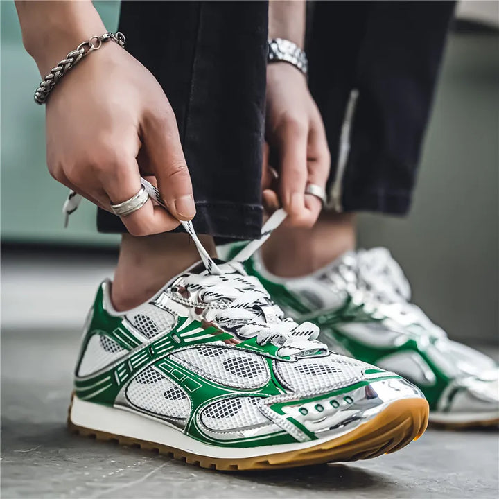 A person wearing rings and a bracelet ties the laces of shiny silver and green lightweight mesh shoes with white soles, cushioned insole, and non-slip outsole, standing on a gray floor.