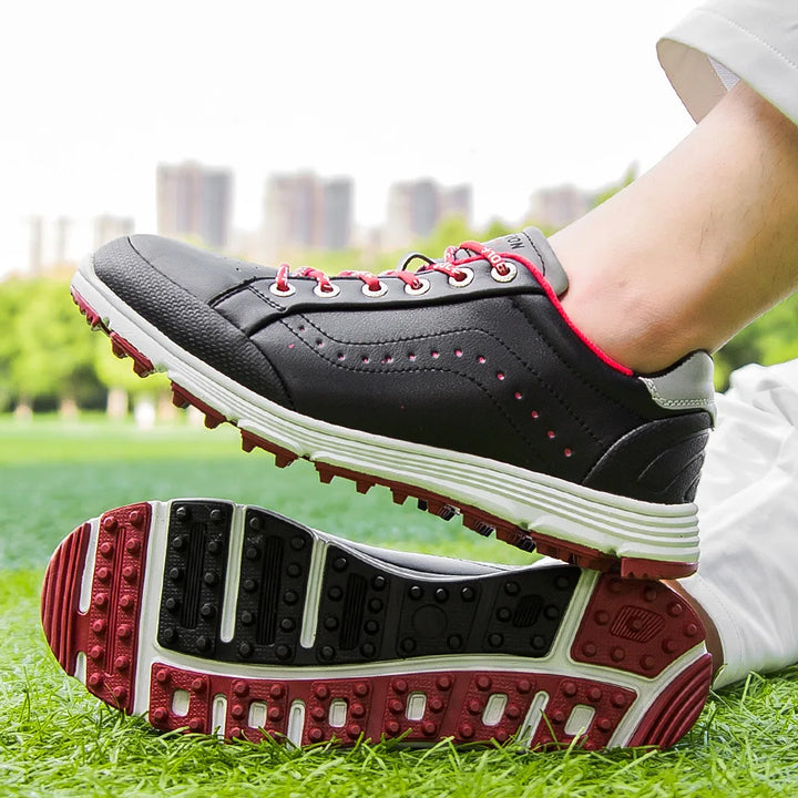 A person wearing black waterproof golf shoes with red accents and white soles stands on green grass, one foot atop the other, showing the shoe’s tread pattern. A city skyline is visible in the blurred background.