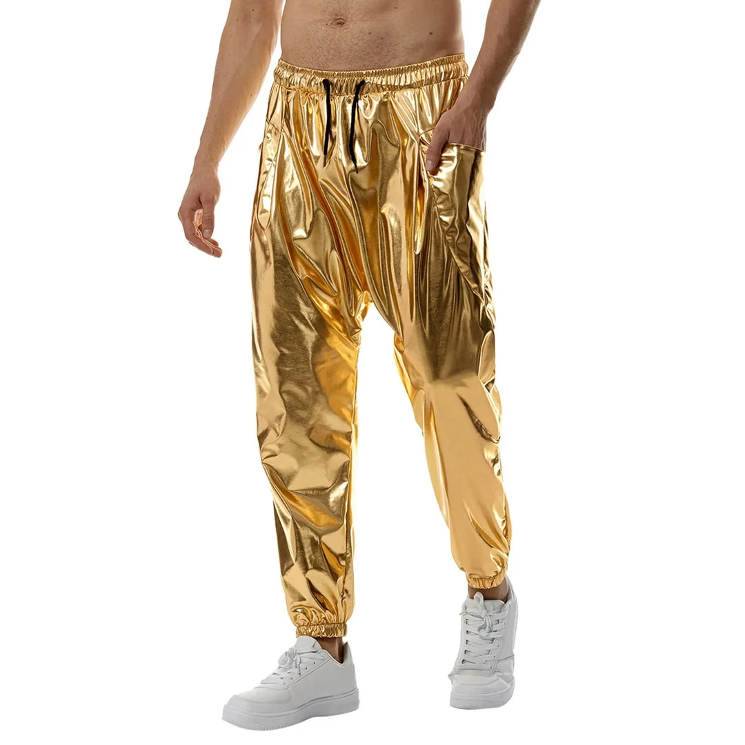 A man wearing shiny gold jogger pants made from lightweight metallic fabric with an elastic waistband and tapered leg, paired with white sneakers. Hes standing against a plain white background, his upper body bare and left hand in his pocket.