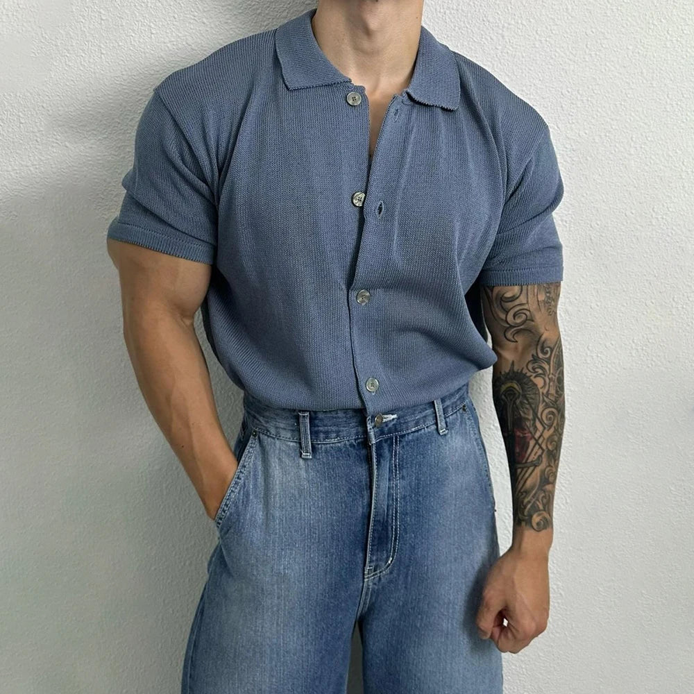 A person wearing a short-sleeve, unisex top and high-waisted blue jeans stands against a white wall, with one hand in their pocket. Their right arm features a large tattoo. Their face is not visible.
