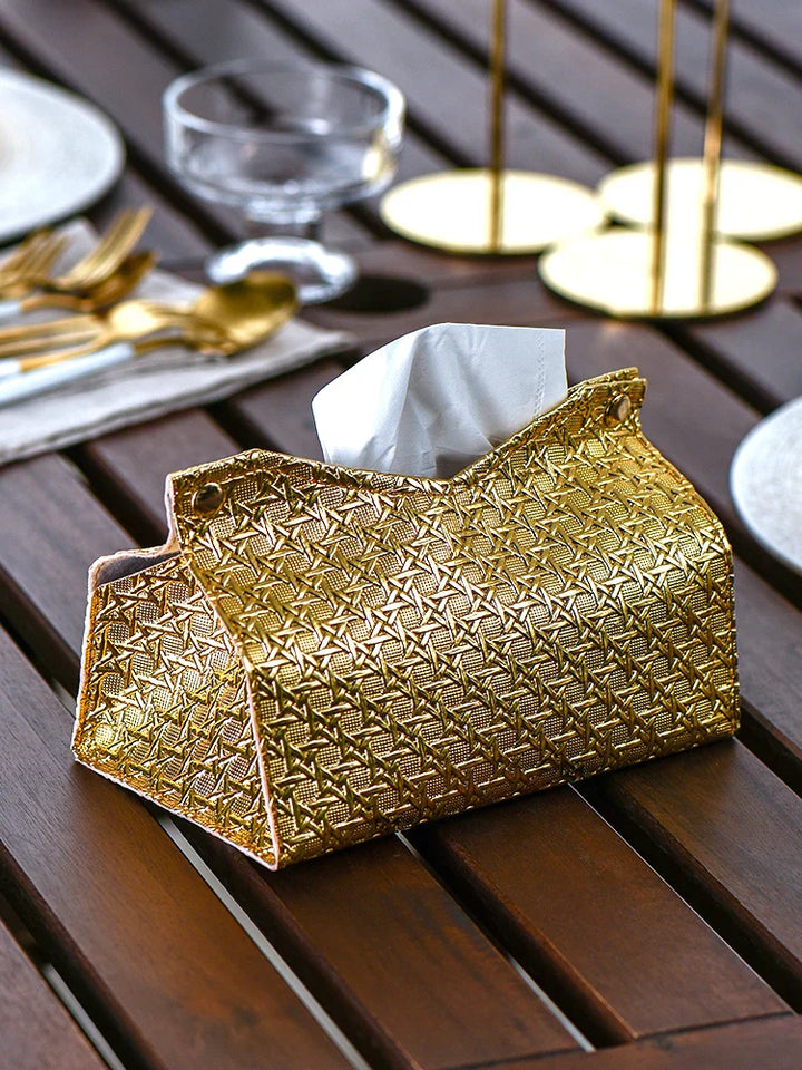 A gold, geometric tissue box adds a touch of luxury to the wooden table, surrounded by gold cutlery, plates, and glassware in an elegant dining setting.