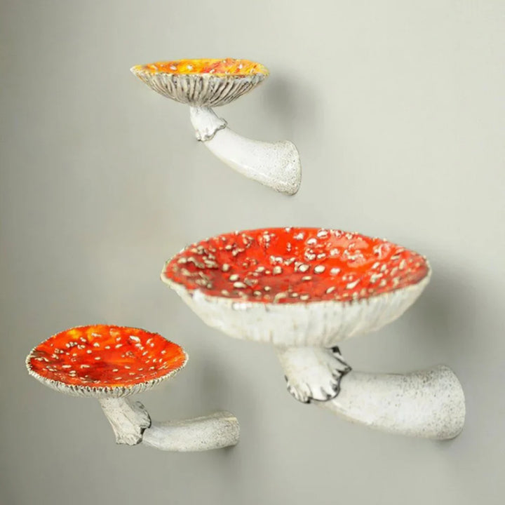 Three wall-mounted ceramic mushroom floating shelves, featuring red and white speckled caps and white stems, create unique home decor with a naturalistic design as they’re arranged diagonally against a light-colored wall.