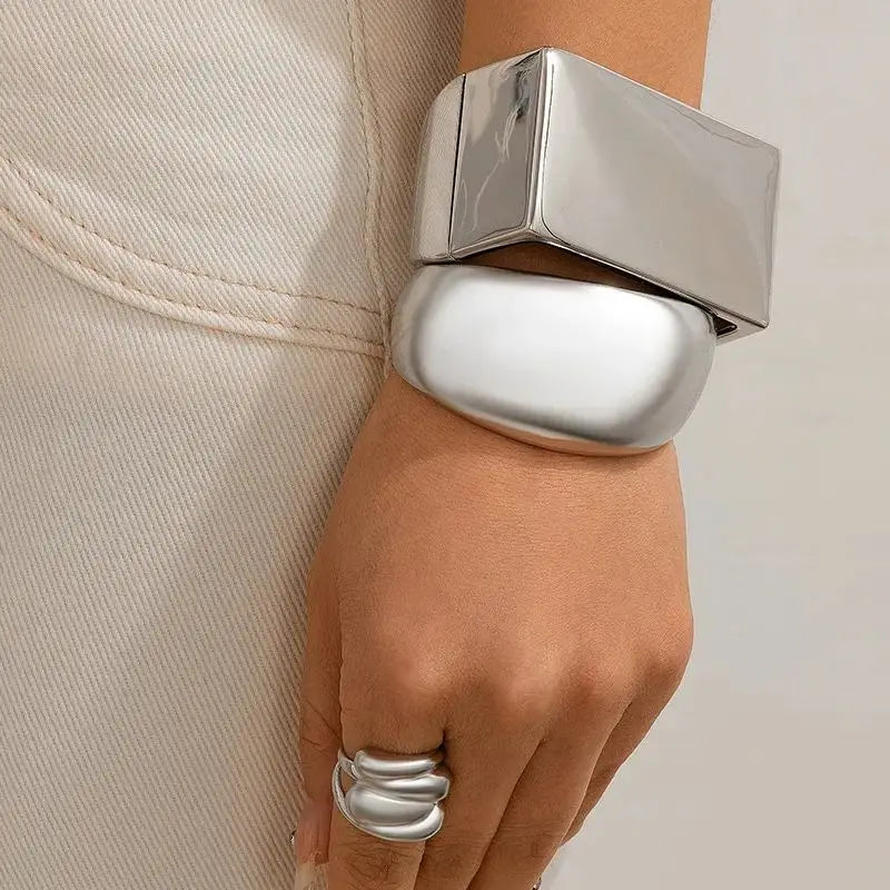 A hand wearing two chunky silver bracelets—one rectangular and one oval-shaped—with a matching silver ring, resting against light-colored pants with visible stitching, showcases unisex jewelry with a bold geometric design.