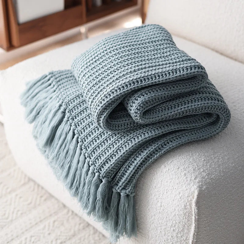 A light blue chunky weave throw with fringed edges is neatly folded and draped over the arm of a white textured sofa.