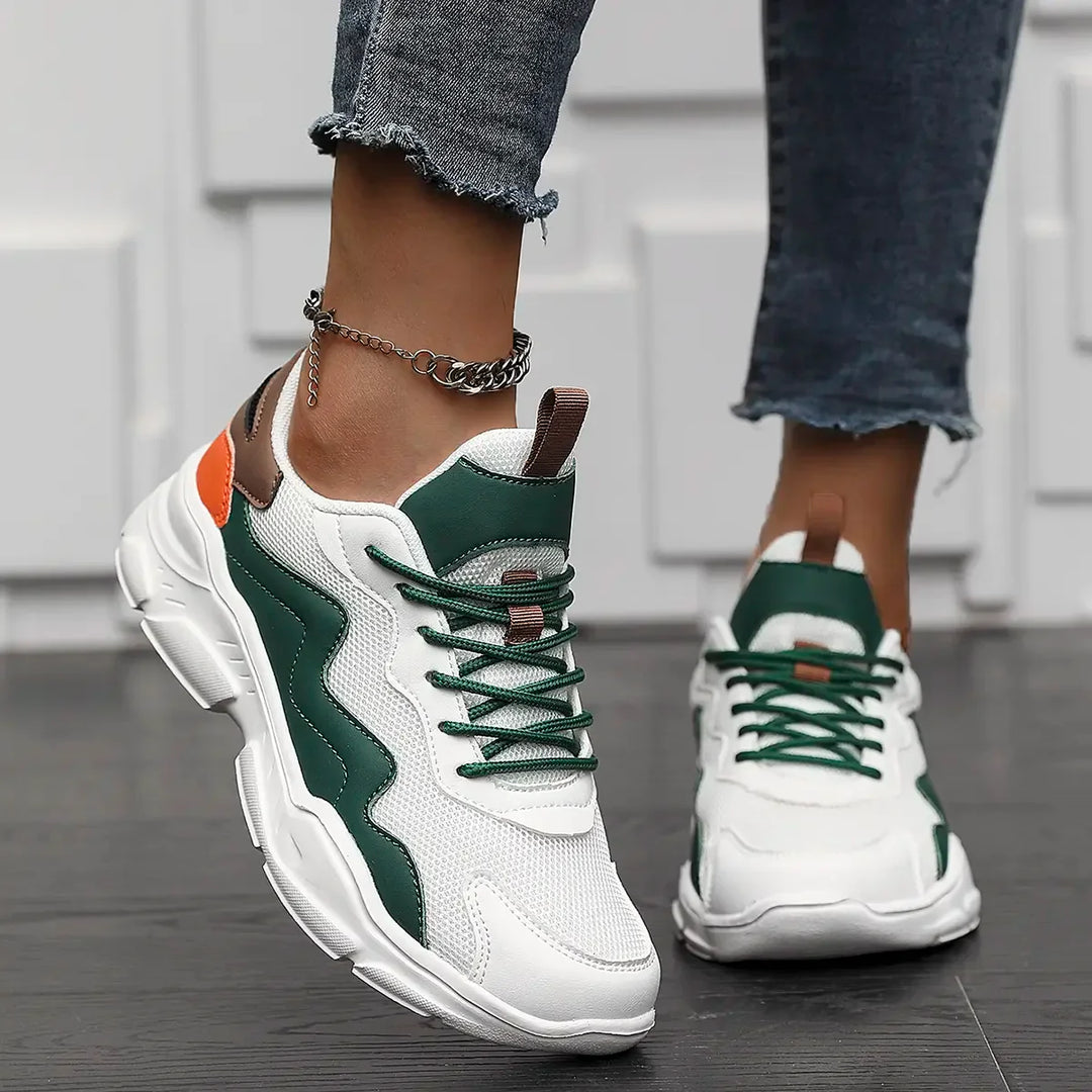 A person wearing lightweight mesh white sneakers with green and orange accents and frayed-hem jeans stands on a dark wood floor. A silver anklet is visible on one ankle, set against a white geometric wall in the background.