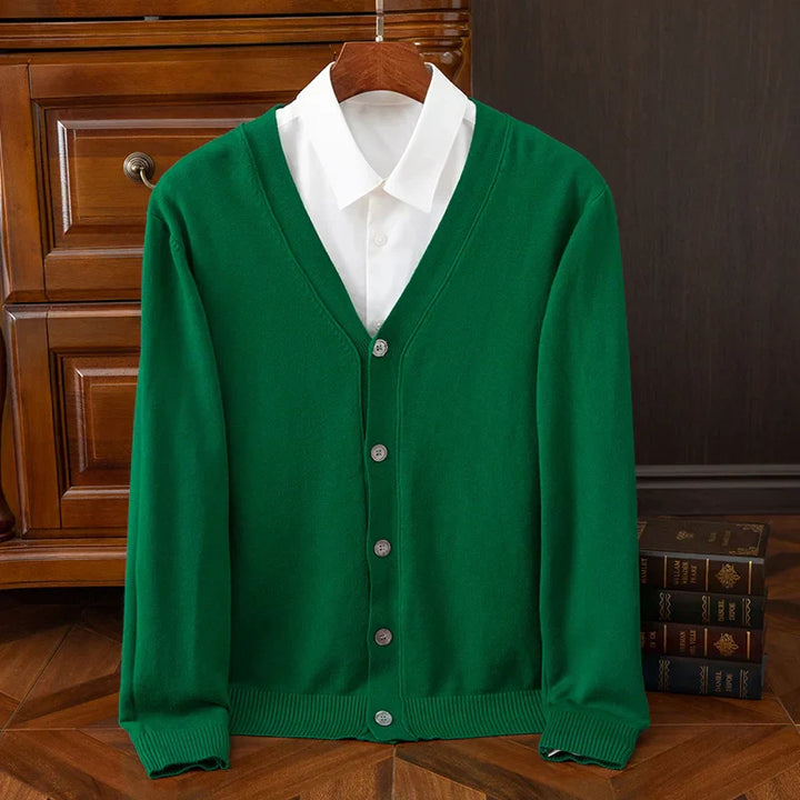 A green relaxed fit button-up cardigan with a V-neckline, layered over a white collared shirt, is neatly hung on a wooden hanger in front of a wooden cabinet and stacked books on a wooden floor.