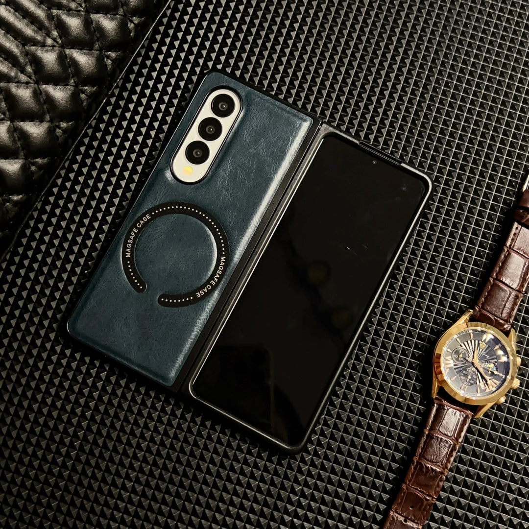 A foldable smartphone in a Samsung Galaxy Z Fold case lies open on a textured black surface, next to a wristwatch with a brown leather strap and gold case. Part of a quilted black item is visible in the top left corner.