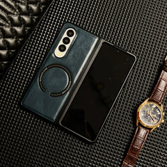 A foldable smartphone in a Samsung Galaxy Z Fold case lies open on a textured black surface, next to a wristwatch with a brown leather strap and gold case. Part of a quilted black item is visible in the top left corner.