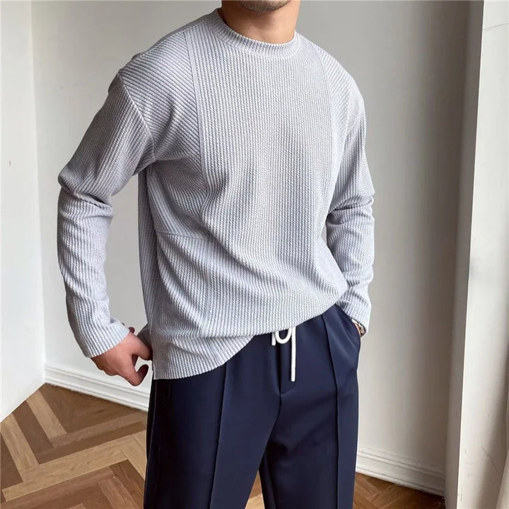 A person stands indoors wearing a light gray Grayson Crew Classic ribbed knit long-sleeve shirt and loose-fitting dark blue trousers with a visible white drawstring, with one hand in their pocket.