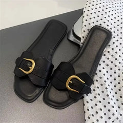 A pair of black leather slides with gold buckles are placed on a dark surface next to a white polka-dotted fabric.