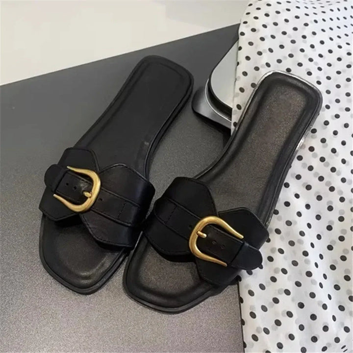 A pair of black leather slides with gold buckles are placed on a dark surface next to a white polka-dotted fabric.
