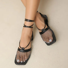 A pair of feet wearing black, square-toed mesh flats with ankle straps. The shoes feature a mesh upper and netted design, offering style and comfort, and are worn on light-colored flooring.
