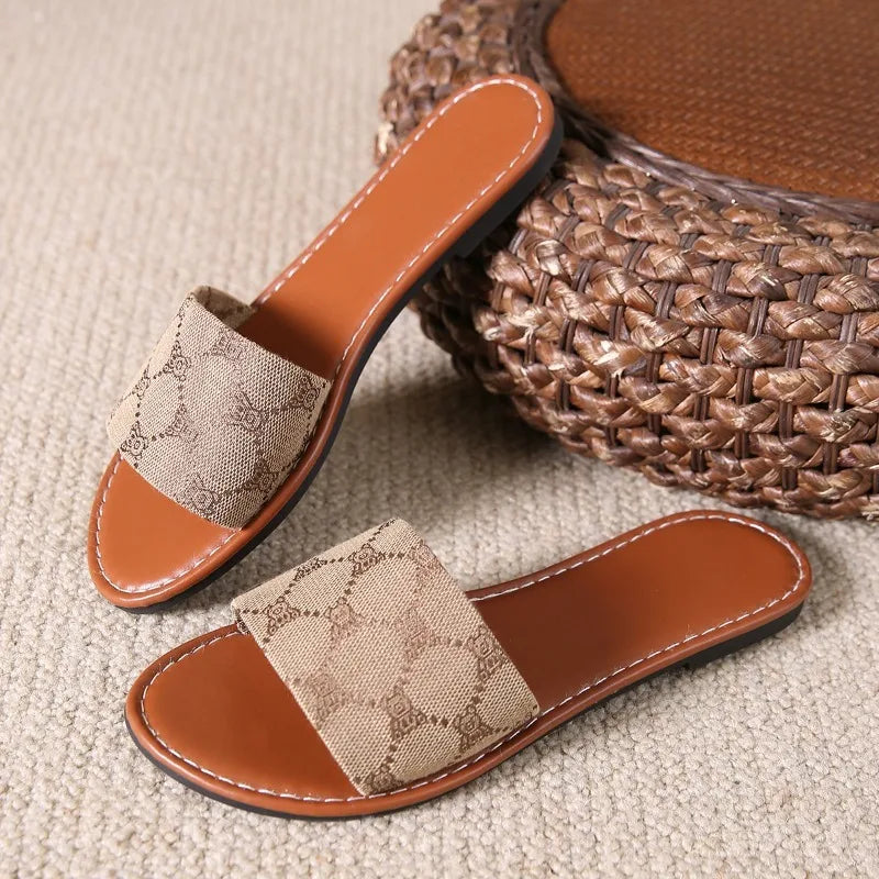 A pair of brown flat sandals with tan fabric straps featuring a subtle pattern, placed on a beige textured carpet next to a woven round ottoman—perfect modern slippers for easy summer style.