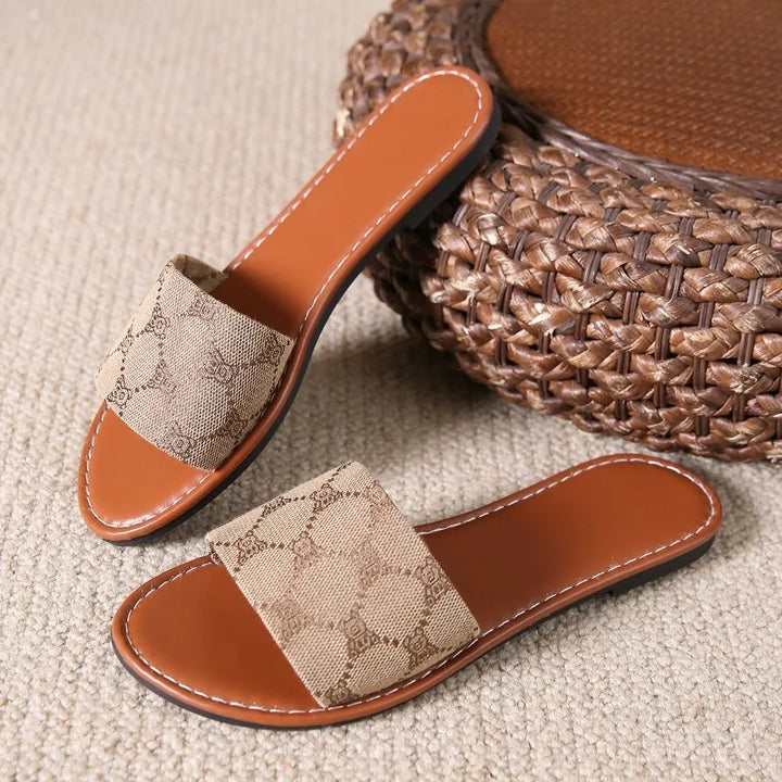 A pair of brown flat sandals with tan fabric straps featuring a subtle pattern, placed on a beige textured carpet next to a woven round ottoman—perfect modern slippers for easy summer style.