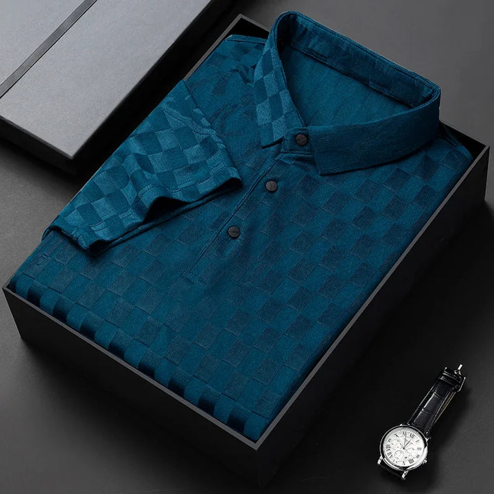 A dark teal, short-sleeve, tailored fit button-up shirt with a checkerboard pattern is neatly folded in a black gift box. A classic wristwatch with a black strap is placed nearby on a black surface.