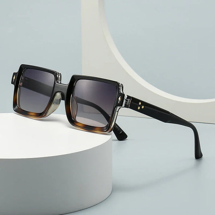 Square-shaped retro sunglasses with thick, dark brown and black gradient frames and gray-tinted lenses offer UV400 protection, displayed on a white, round platform against a minimalist, light-colored background.