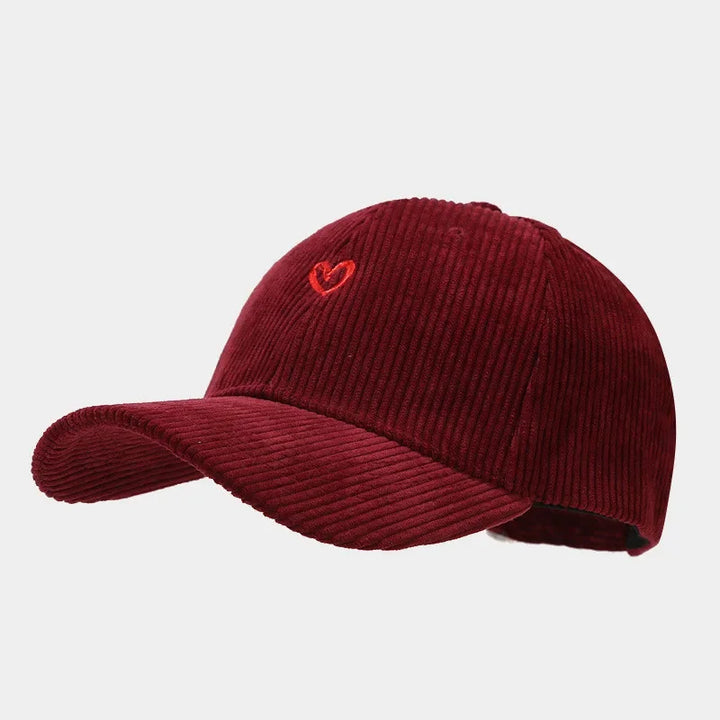 A burgundy corduroy baseball cap featuring a small heart embroidery on the front, complete with a FlexFit band for added comfort. Shown against a plain light gray background.