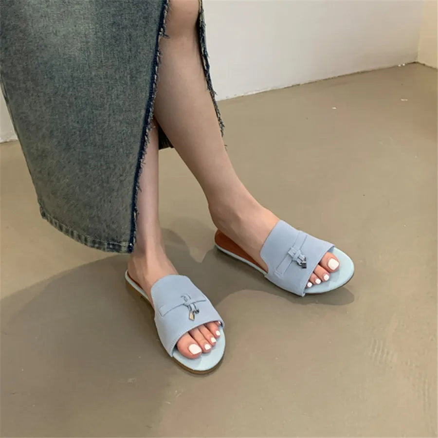 A person wearing a long, frayed denim skirt sits with legs crossed, showing light blue slide sandals crafted with a cushioned insole and decorative bows, highlighting freshly pedicured toenails on a smooth beige floor.