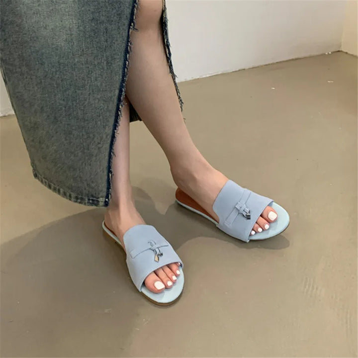 A person wearing a long, frayed denim skirt sits with legs crossed, showing light blue slide sandals crafted with a cushioned insole and decorative bows, highlighting freshly pedicured toenails on a smooth beige floor.