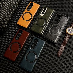 Five leather cases, including a Samsung Galaxy Z Fold case, in brown, green, black, burgundy, and blue are displayed on a textured black surface beside a brown leather wristwatch and a partially visible quilted black object.