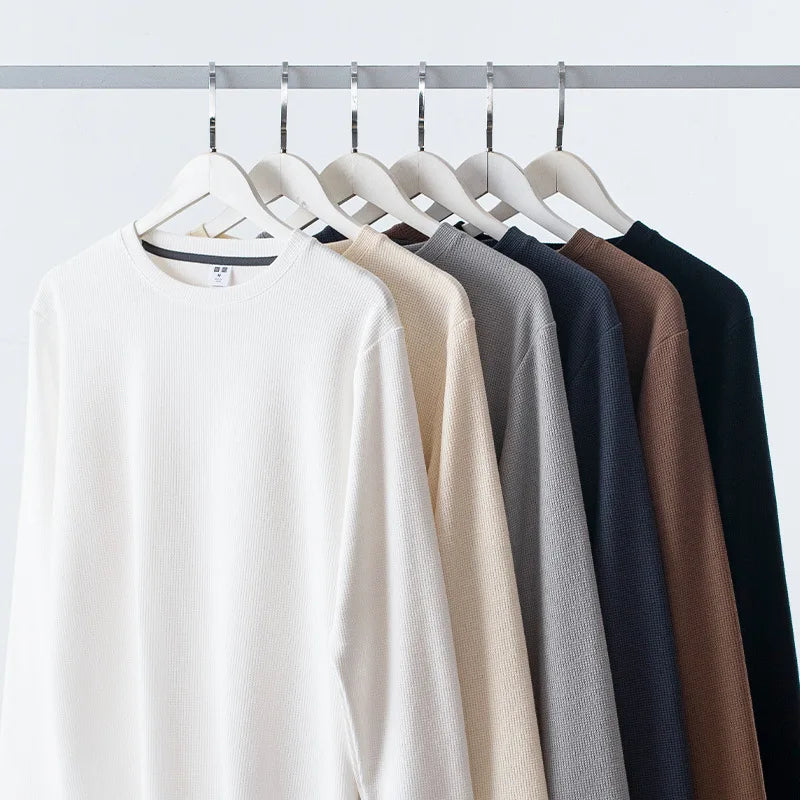 Six heavyweight long-sleeve shirts in white, beige, light gray, gray, brown, and black made from premium cotton hang neatly on white hangers on a metal clothing rack against a plain white background.