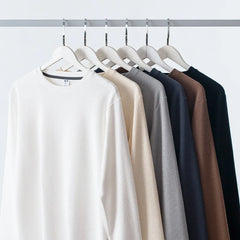 Six heavyweight long-sleeve shirts in white, beige, light gray, gray, brown, and black made from premium cotton hang neatly on white hangers on a metal clothing rack against a plain white background.