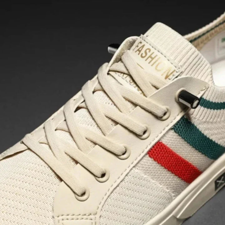 Close-up of a beige sneaker with beige laces, ribbed tongue, and FASHION label. The side features green and red stripes on the breathable mesh upper. The shoe is set against a dark background.