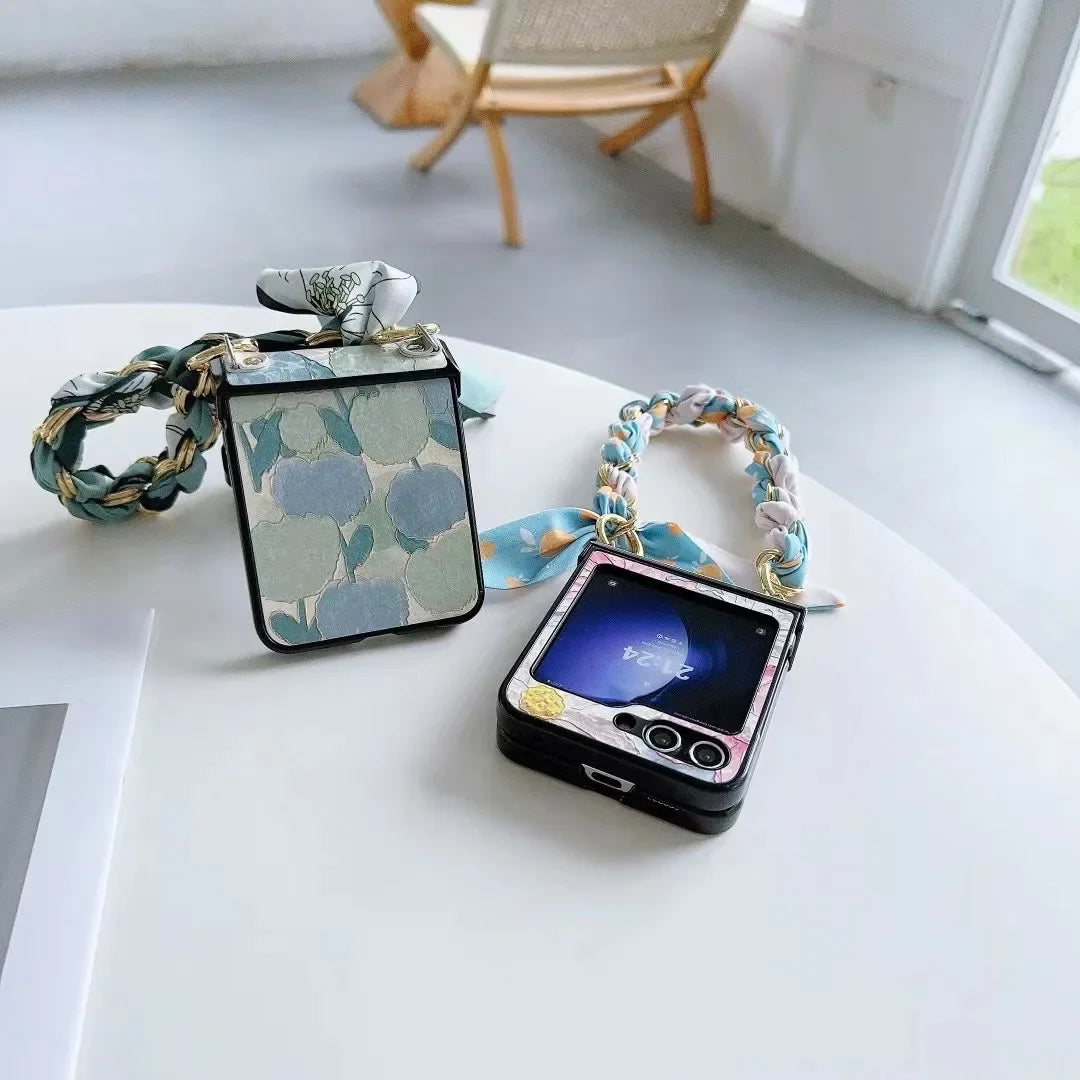 Two smartphones with patterned cases and chain straps rest on a white table in a minimal, bright room with a chair and window in the background. One phone screen is on, showing a blue wallpaper and highlighting the slim design.