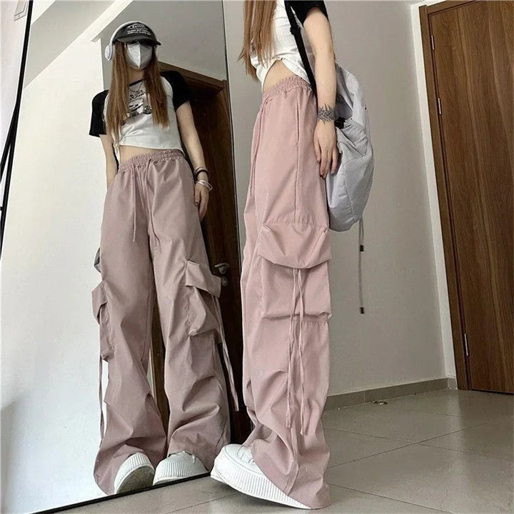 A person in high-waist pink cargo pants with cargo pockets, a black and white crop top, white sneakers, a baseball cap, a face mask, and a backpack stands before a mirror in a minimal room with tiled floor.