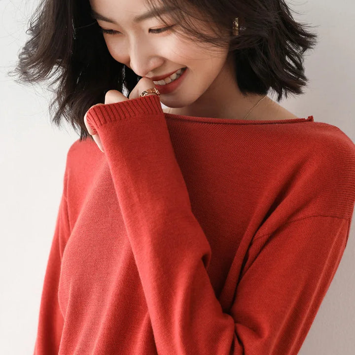 A woman with shoulder-length dark hair wears a red, relaxed fit sweater. She smiles softly, looking down, with one hand touching her face as if shy or playful. The background is plain and light-colored.