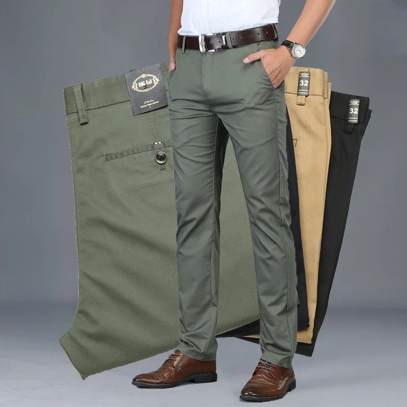 A man wearing olive green dress pants with a tailored fit, brown belt, white shirt, and brown shoes stands in front of three pairs of folded pants in olive green, beige, and black—highlighting their versatile style against a plain grey background.